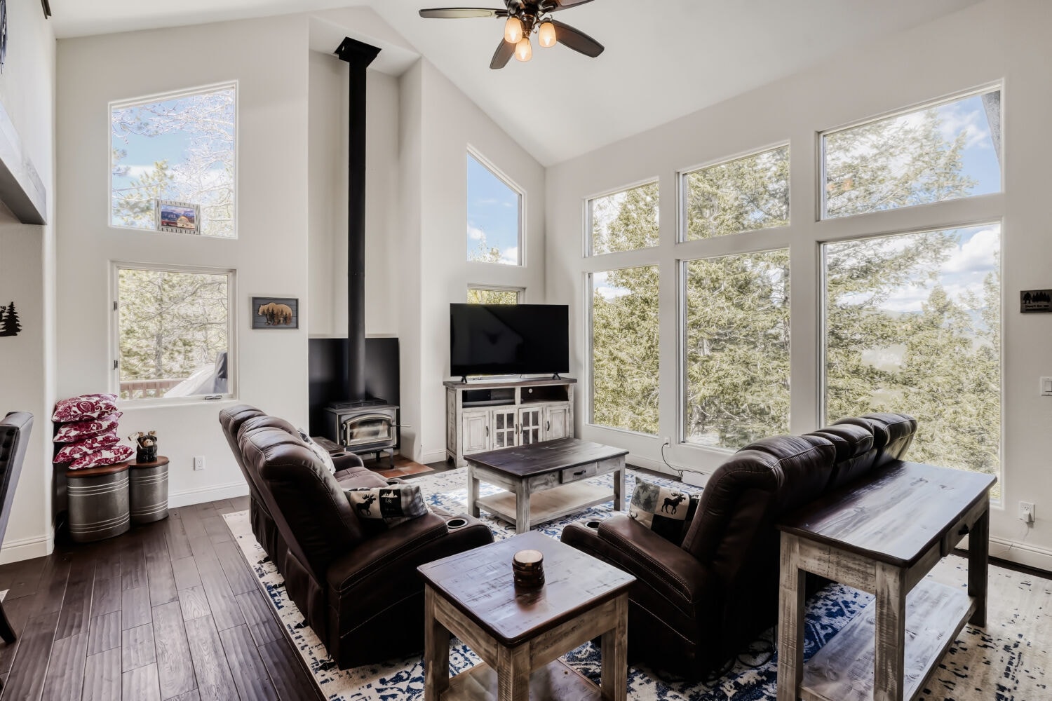 Living room with cathedral ceilings, wood stove, and wall-to-wall windows framing the pines